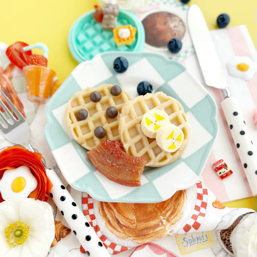 Breakfast scene with waffles, bacon, and fruit on a checkered plate with a yellow background.