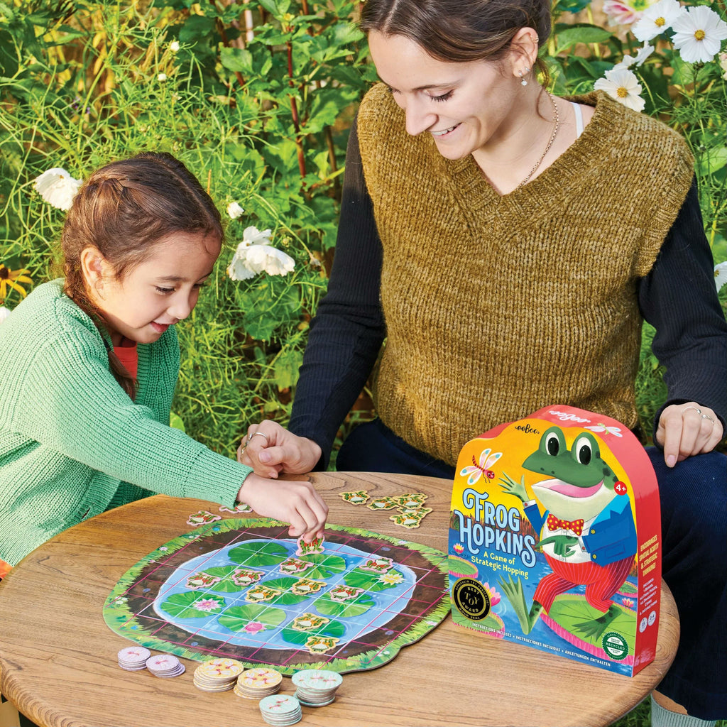 Woman and child playing a board game outdoors with 'Frog Hopkins' packaging visible.