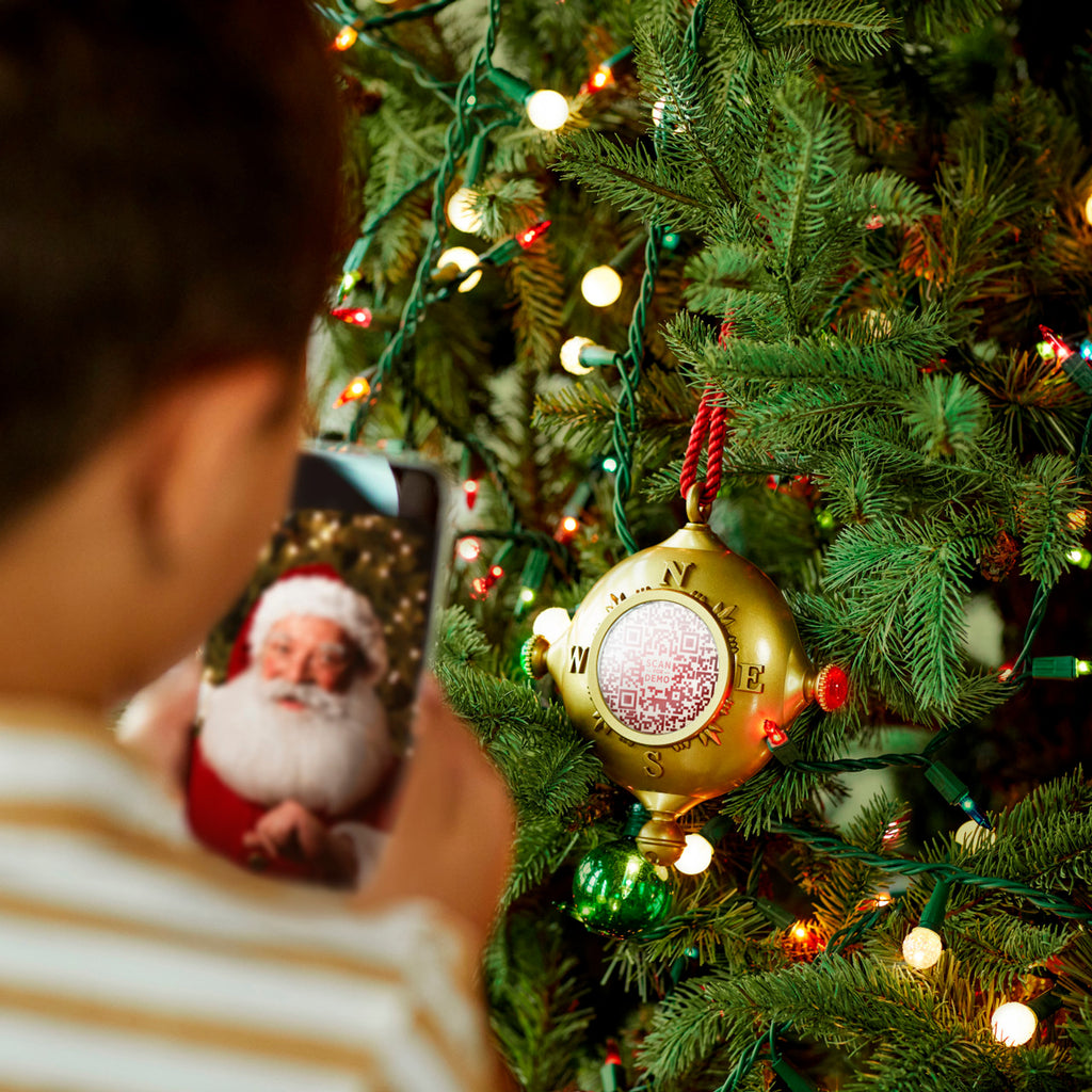 Image of child holding phone and viewing a video of Santa.  In the background is a Christmas tree with Santa's Kindness ornament hanging on it.